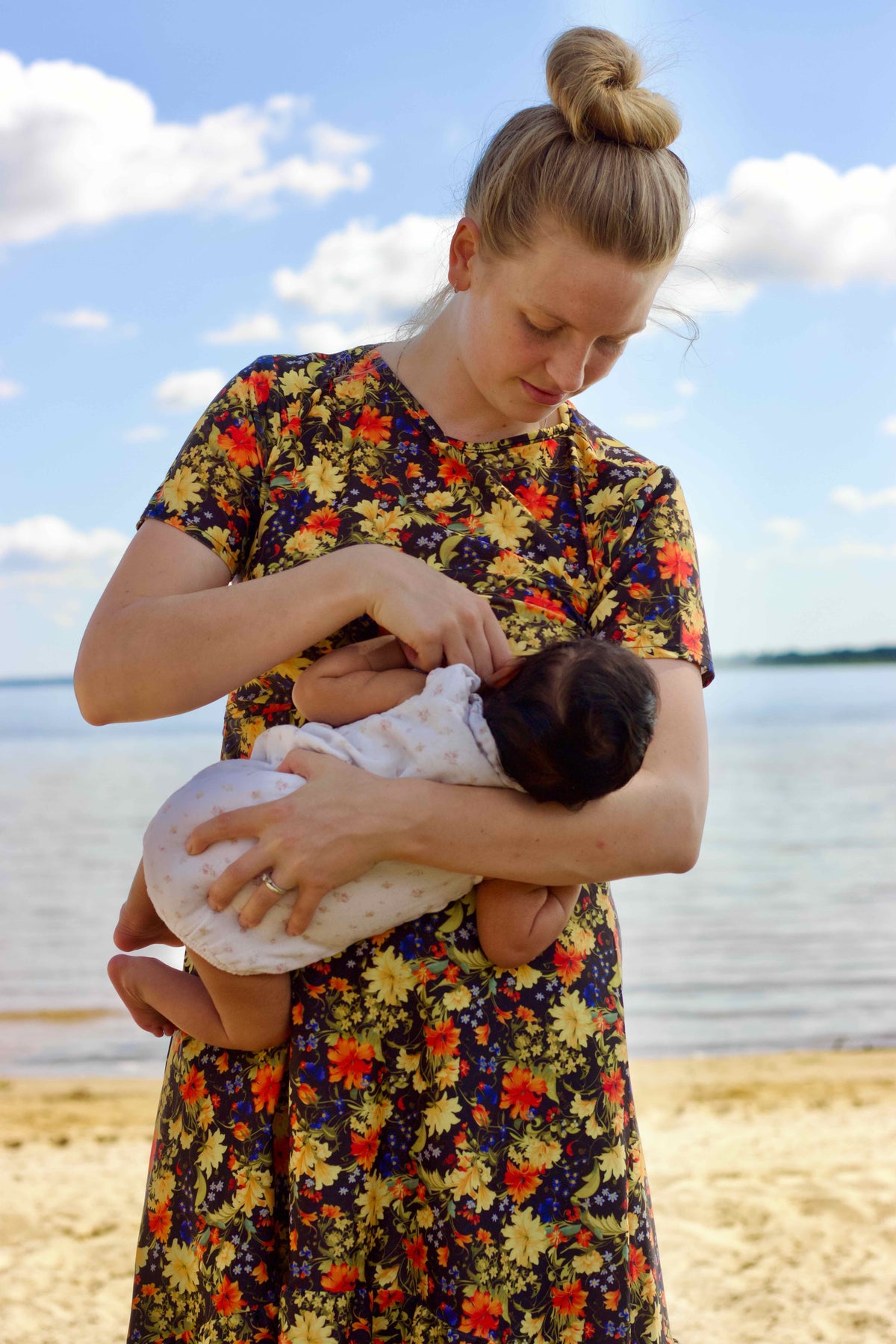 A woman in a colorful floral swimdress holds a swaddled baby on a sandy beach with water and a blue sky in the background. showing the discreet nursing access