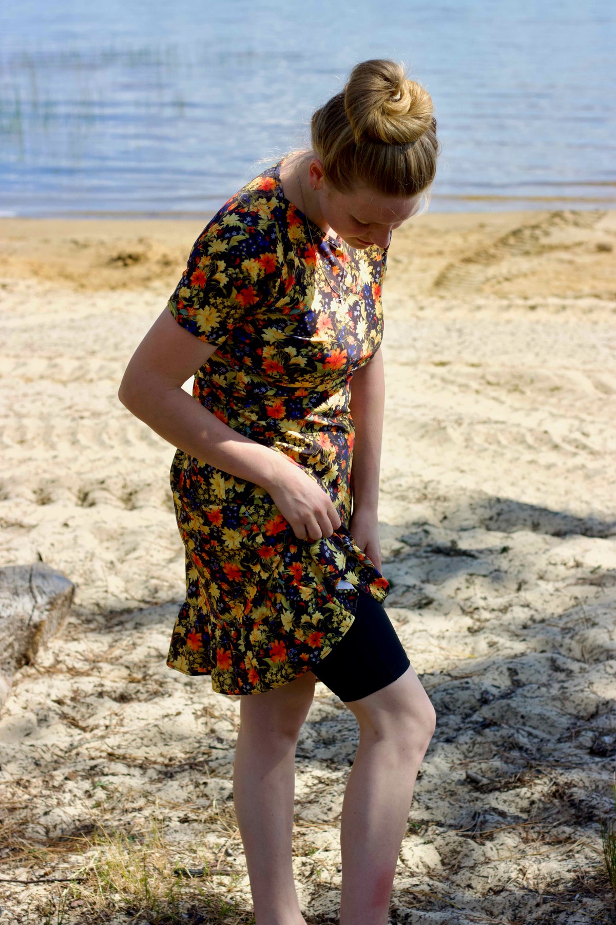 A woman standing on a sandy beach near the water, wearing a short-sleeve modest swimwear over black swim shorts.