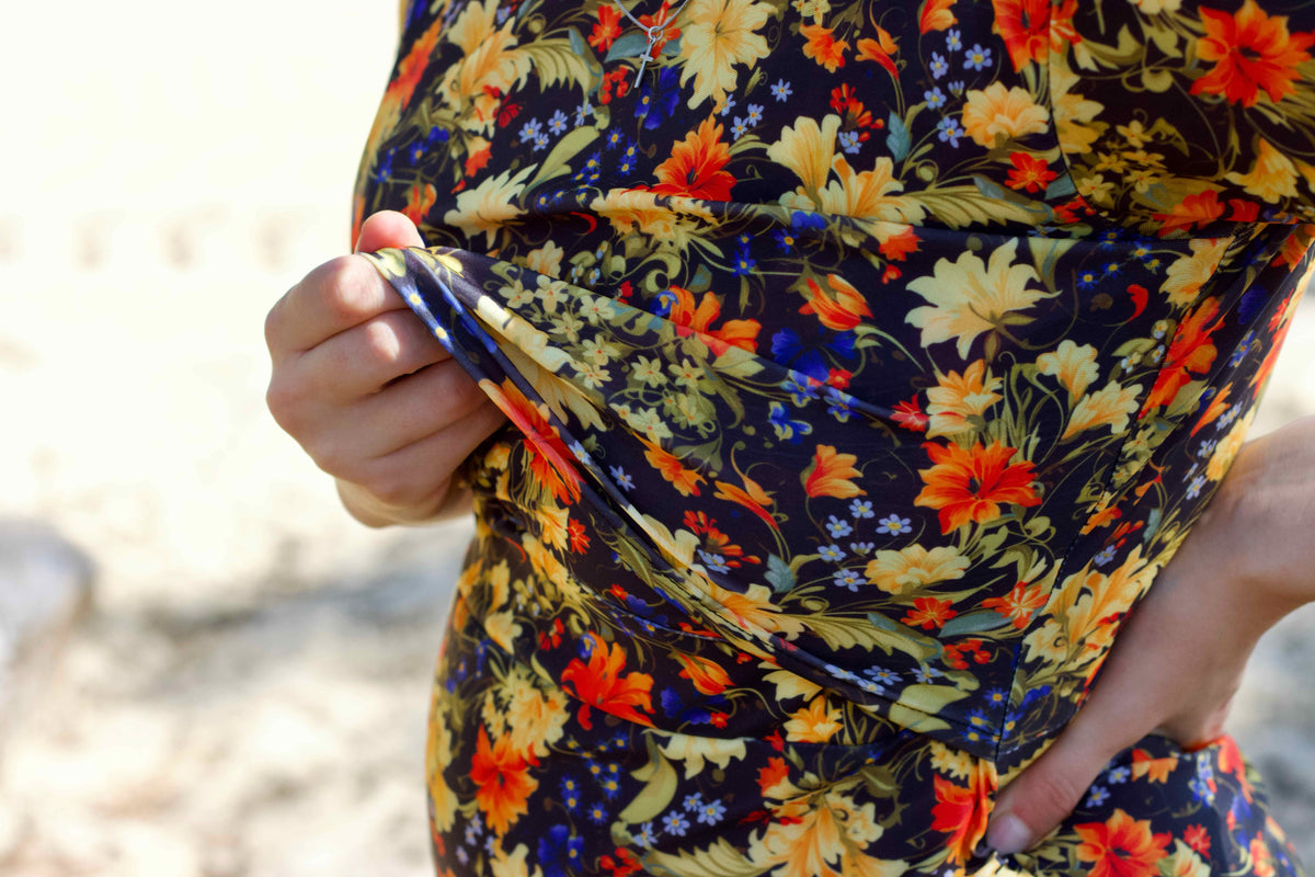 Close-up of a lady wearing a dark floral nursing friendly swimdress or wrap with yellow, orange, and blue flowers, holding the fabric at the waist with one hand showing the nursing access.