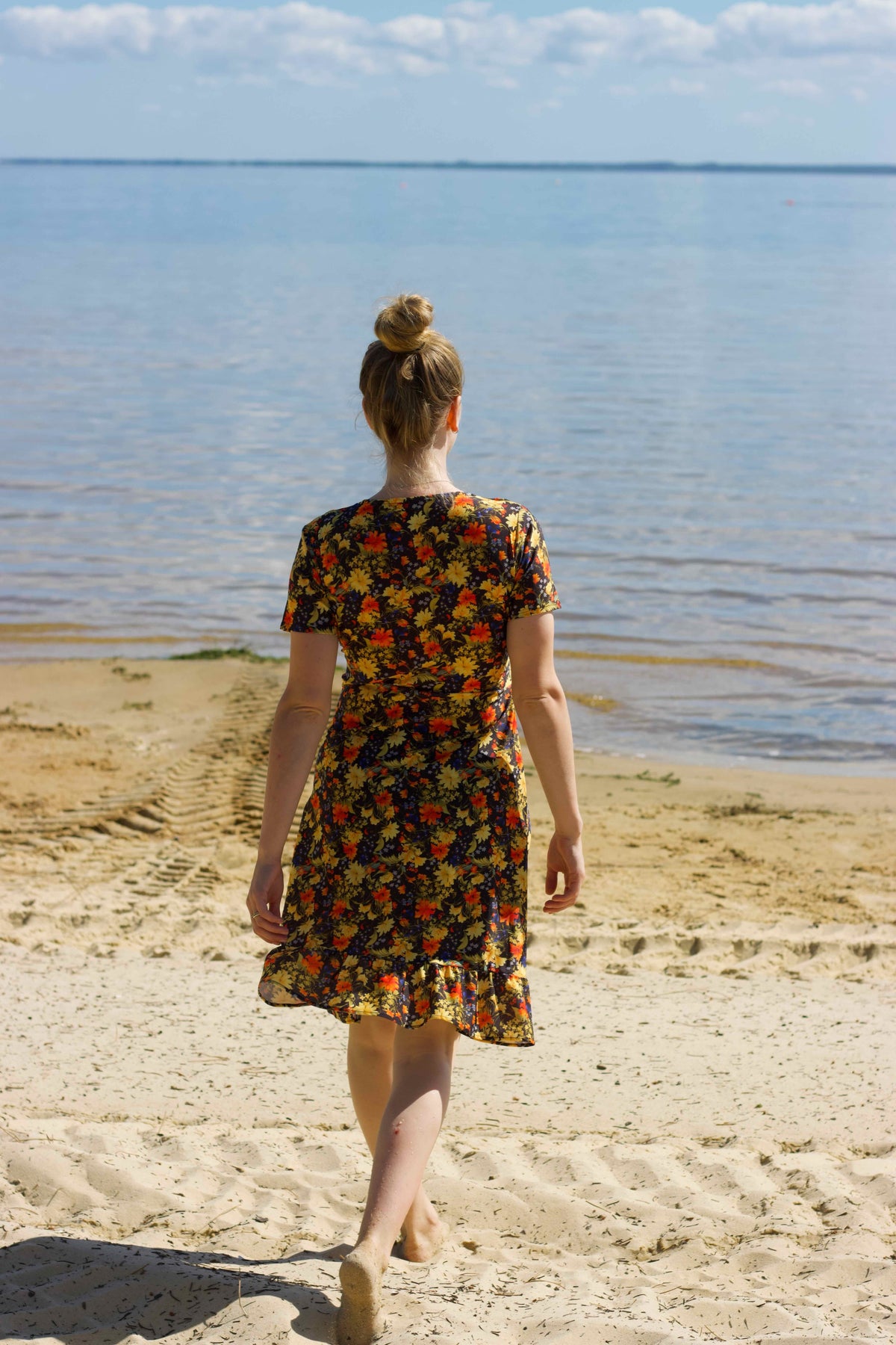 A woman seen from behind walking on a sandy beach toward the water, wearing a short-sleeve swimdress with full coved  back and knee length skirt with a multicolor print on a dark background.
