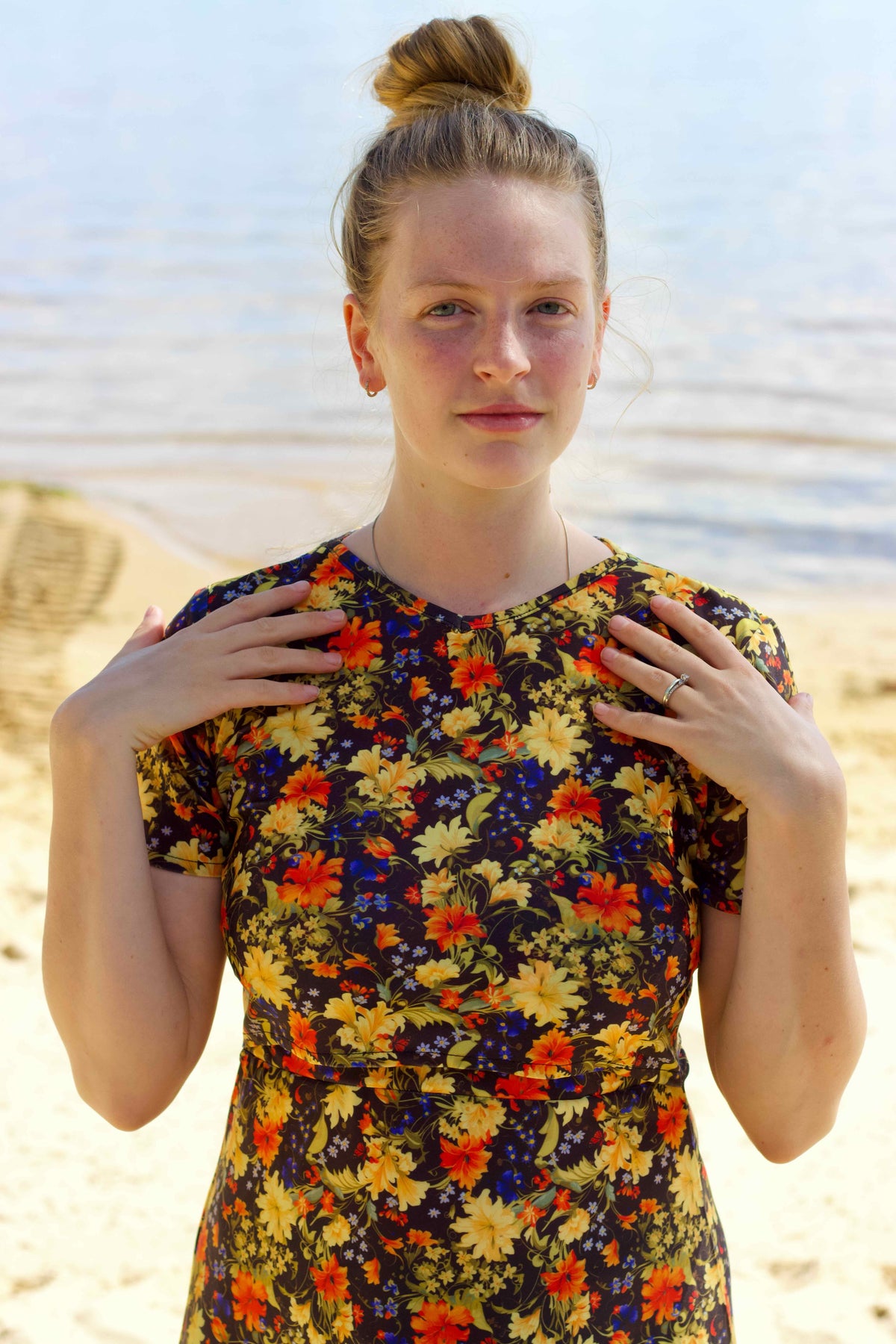A woman with her hair in a bun stands on a sandy beach with water in the background, wearing a short-sleeve modest high neck swimdress with a dense multicolor floral print on a dark base.
