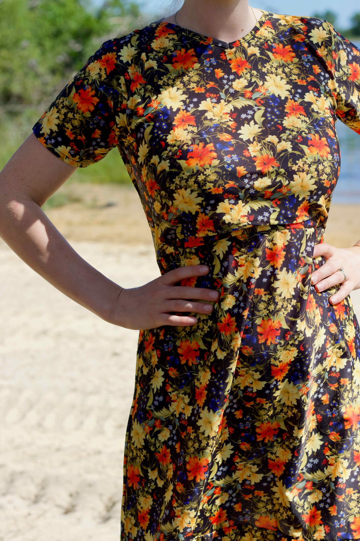 A woman wearing a short-sleeve V-neck christian swim dress with a dense floral print in yellow, orange, and red on a dark navy background, standing outdoors on a sandy beach with green foliage in the background.