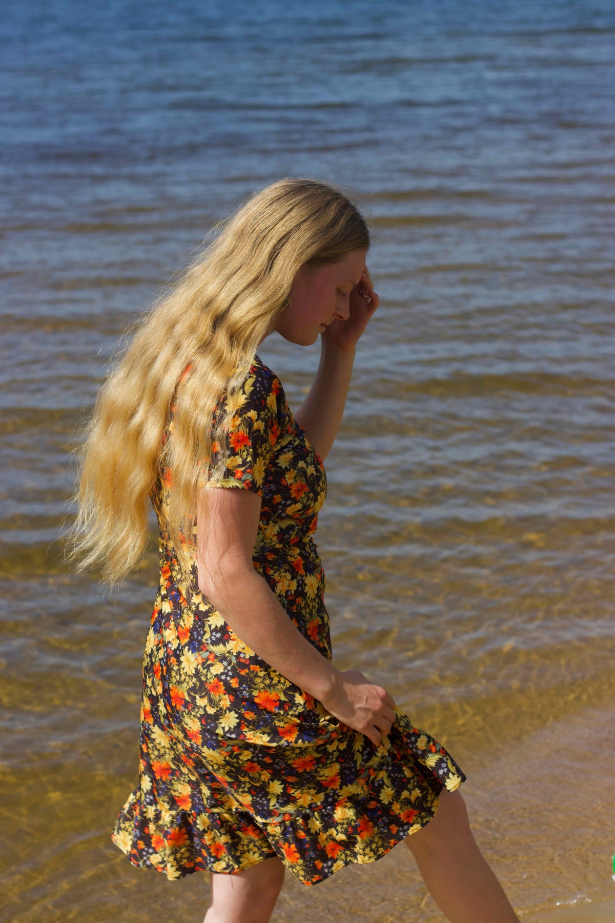 A woman with long blonde hair stands at the edge of a body of water wearing a short-sleeved black swim dress with a vibrant yellow, orange, and red floral pattern.